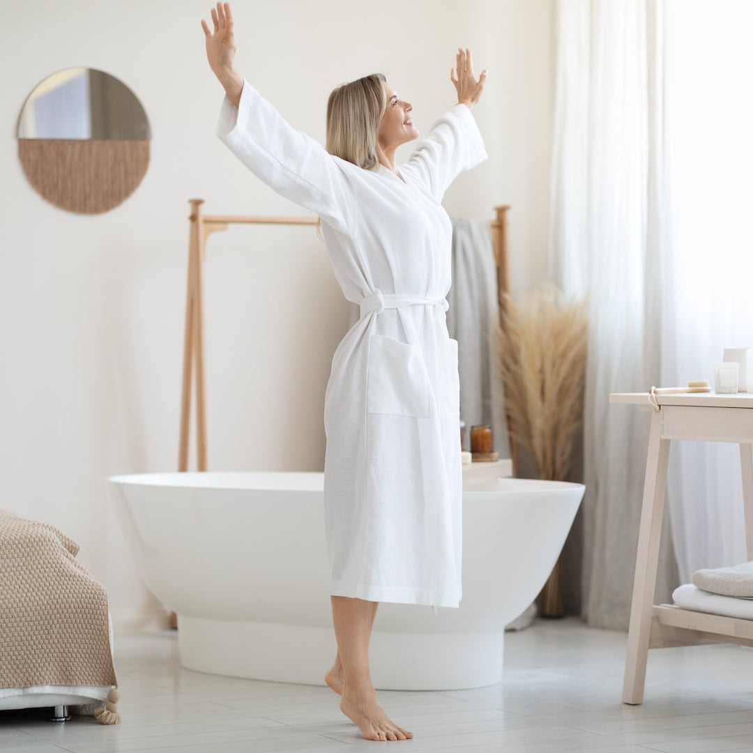 Woman in a white Harbourloom bathrobe standing in a sunlit bathroom, embodying the calm elegance of the Harbourloom Bath collection.