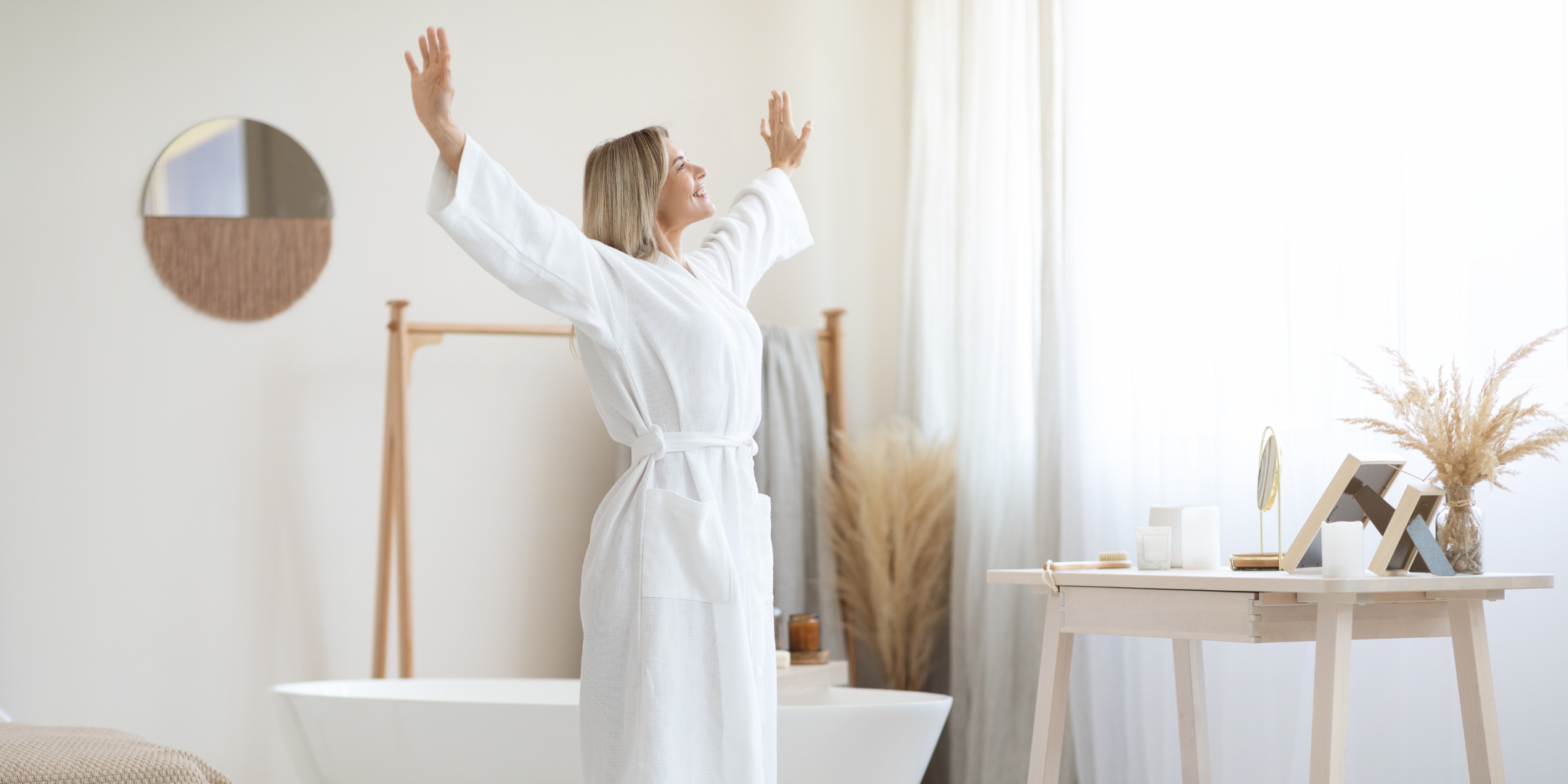 Woman in a white bathrobe stretching in a bathroom with a bathtub and wooden table.