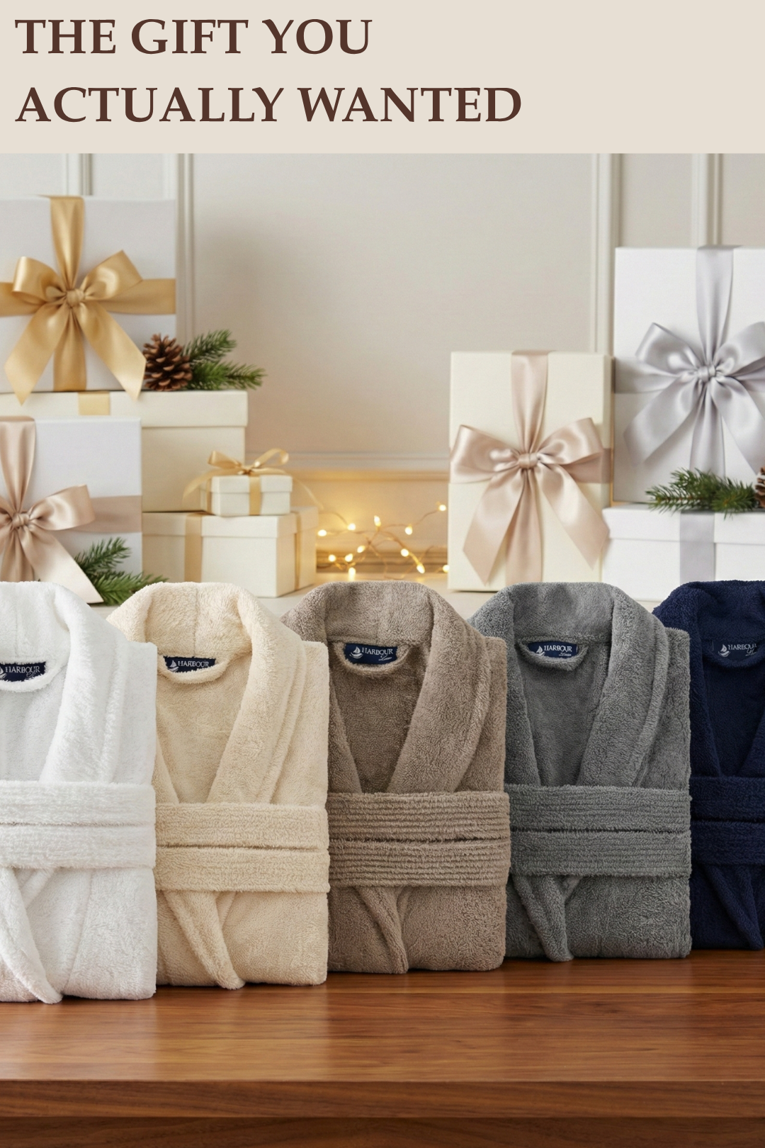 Five premium Harbour Loom Turkish cotton spa bathrobes neatly folded on a wooden table, available in white, cream, taupe brown, charcoal grey, and navy blue, with elegant holiday gift boxes in the background